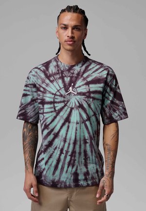 Mand med fletninger og tatoveringer på armene bærer en lilla og turkis spiral tie-dye t-shirt med en hvid Jumpman-logo.