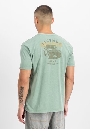 Mann mit kurzem Haar, Tätowierungen an den Armen, trägt ein verblasstes grünes T-Shirt mit einem Vintage-Lkw und den Texten "BL LEGENDS" und "ALPHA INDUSTRIES" auf der Rückseite.