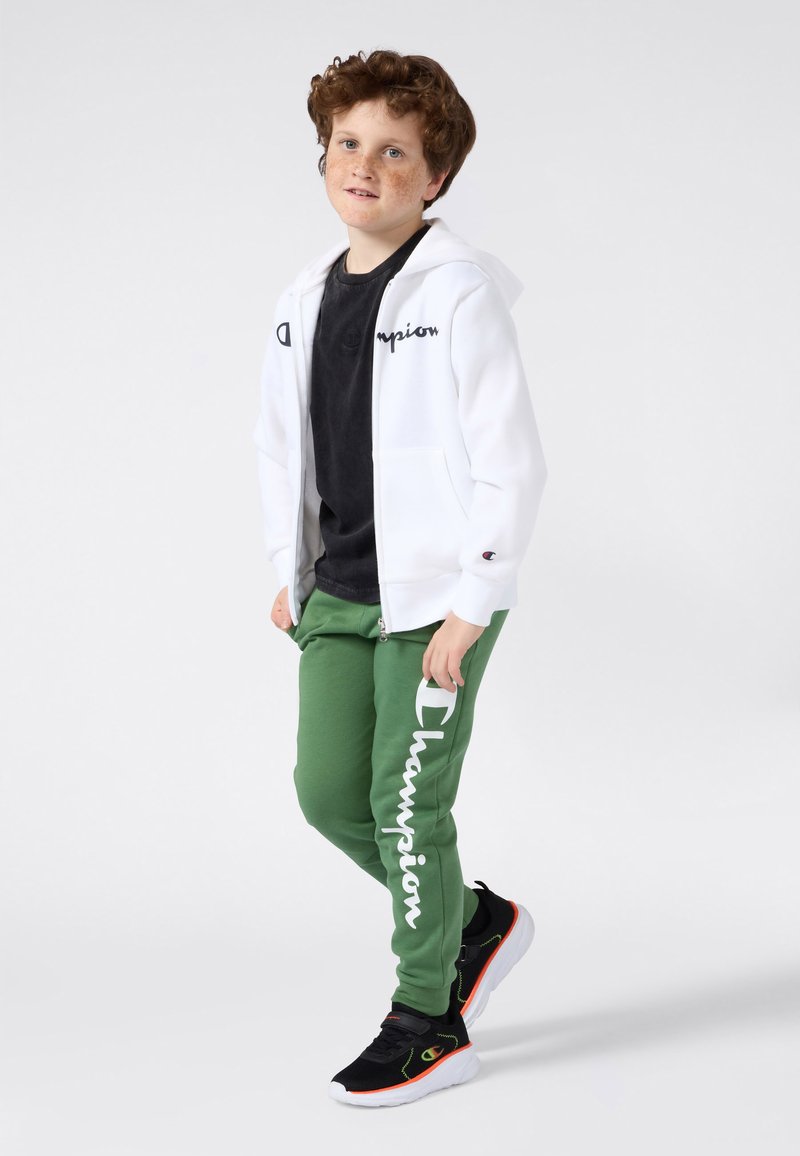 Witte rits hoodie met "Champion" logo, zwarte crewneck shirt, groene trainingsbroek en zwarte sneakers met neon-accenten.