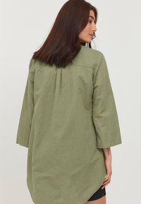 Camicia leggera verde oliva con maniche a tre quarti, orlo asimmetrico e texture sottile. Presenta una yoke sul retro e un dettaglio a pieghe.