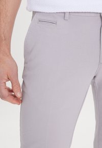 Light gray trousers made from textured fabric, featuring a slim fit, side pockets, and a smooth waistband. The hand is positioned near the seam.
