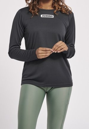 Persona con una camiseta negra de manga larga de Hummel y leggings verdes, de pie con las manos suavemente entrelazadas a la altura de la cintura.