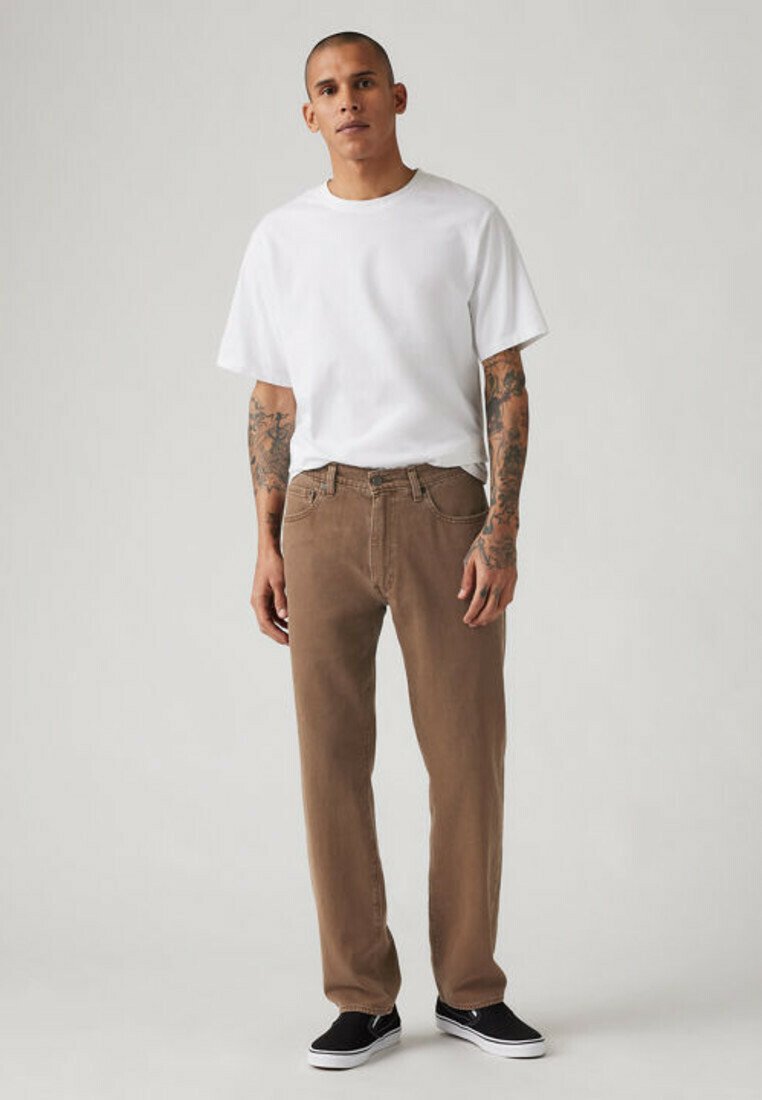 Man with tattoos wears white t-shirt, brown pants, and black slip-on shoes, standing against plain light gray background.