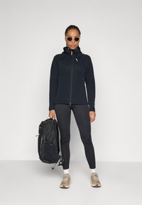 Schwarze Zip-Jacke mit Kapuze, kombiniert mit schwarzen Leggings und beigen Turnschuhen. Das Modell hält einen schwarzen strukturierten Rucksack.