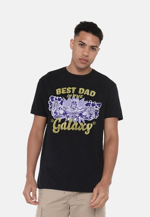 Young man wearing a black T-shirt with a muscular cartoon character and text reading "Best Dad in the Galaxy" in gold and purple colors.