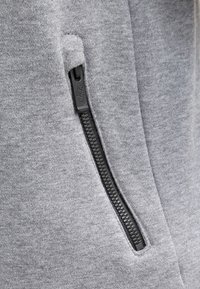 Grauer Sweatshirtstoff mit einer Reißverschlusstasche, die eine strukturierte Oberfläche und einen schwarzen Reißverschluss aufweist. Der Reißverschluss ist mit einem markierten Ziehgriff versehen.