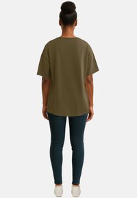 T-shirt verde oversize con maniche corte, texture liscia, scollo rotondo e vestibilità ampia abbinata a jeans skinny blu scuro e sneakers bianche.