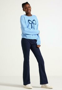 Lyseblå strikket sweater med et navy sløjfedesign, parret med mørkeblå fløjl flared bukser og hvide sneakers.