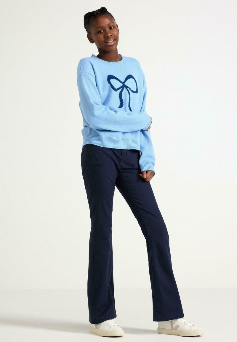 Helles blaues gestricktes Sweatshirt mit einem dunkelblauen Schleifen-Design, kombiniert mit dunkelblauen Corduroy-weit geschnittenen Hosen und weißen Sneakers.