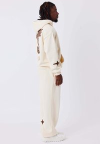 Beige katoenen hoodie met een bruine grafiek op de achterkant en bijpassende joggingsbroek. De broek heeft een bruine sterdetail bij de zoom en wordt gedragen met witte sneakers.