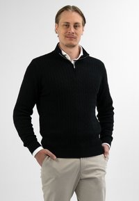 Man staat met handen in zijn zakken, gekleed in een zwart gebreid ritssweater over een wit shirt en een beige broek, tegen een effen achtergrond.