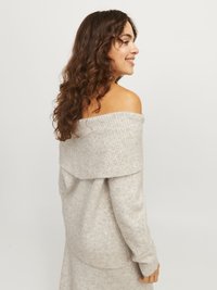Young woman with long curly hair wearing an off-the-shoulder light gray sweater, smiling and looking to her right.