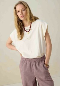 Un haut blanc ample avec des manches retroussées, associé à un pantalon rayé crème et bordeaux ; des colliers en or et en perles bordeaux superposés sont visibles.