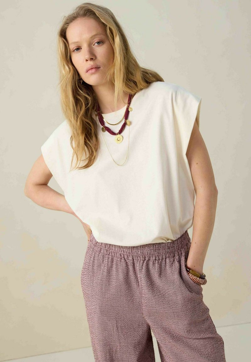 Un haut blanc ample avec des manches retroussées, associé à un pantalon rayé crème et bordeaux ; des colliers en or et en perles bordeaux superposés sont visibles.