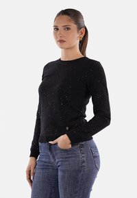Donna con coda di cavallo che indossa un maglione lungo a maniche nere con macchie e jeans blu, in piedi con una mano in tasca su uno sfondo semplice.