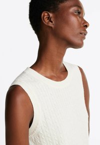 Dzianinowy bezrękawnik w kolorze off-white, z teksturowanym wzorem w warkocze, okrągłym dekoltem i ściągaczem. Gładki materiał i dopasowany krój.