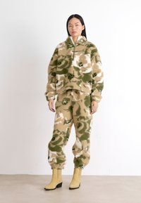 Ensemble en polaire comprenant un pull et un pantalon aux motifs camouflage vert, beige et blanc. Présente un col zippé à moitié et des poignets élastiques.