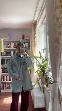 Giacca di denim azzurro oversize con due tasche frontali, chiusura con bottoni e cuciture visibili. Indossata sopra pantaloni a quadri scuri.
