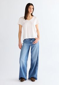 Young woman stands looking sideways, wearing a plain white V-neck T-shirt, wide-leg blue jeans, and brown moccasin shoes.