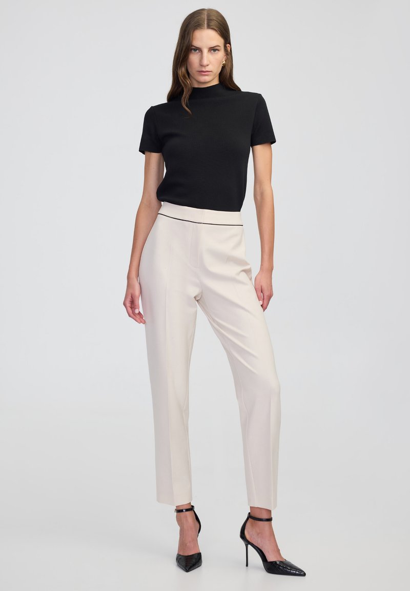 Zwart kortarm shirt met een hoge hals, gecombineerd met beige op maat gemaakte broek en zwarte puntige hakken, met een slim fit en strakke lijnen.