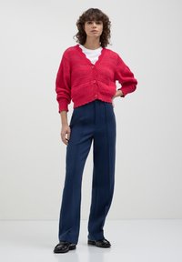 Femme debout en pantalon bleu marine, T-shirt blanc et cardigan boutonnière texturé rose vif, les mains sur les hanches, sur fond uni.