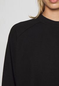 Parte superior del torso de una persona que lleva una camiseta de cuello redondo de manga larga negra, contra un fondo claro y liso.