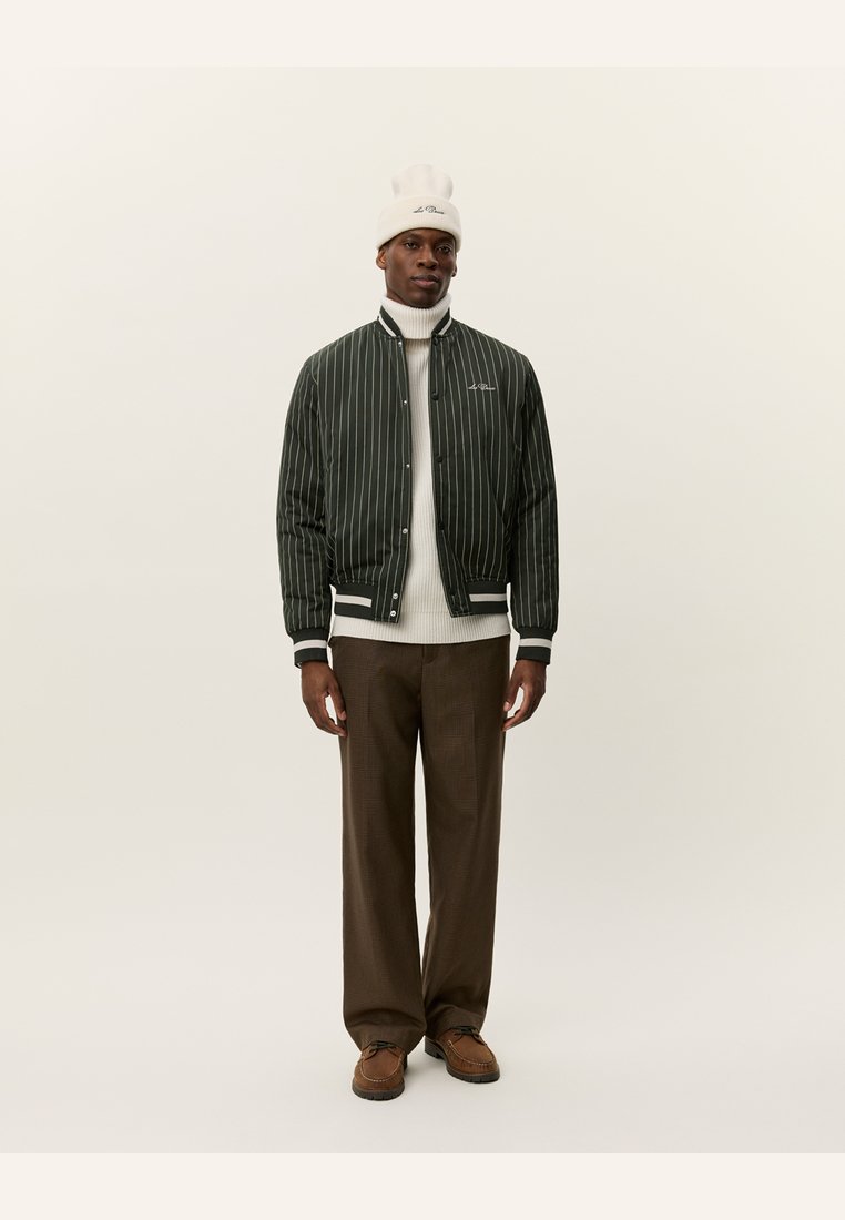 Green striped bomber jacket with a white knit turtleneck, brown plaid trousers, and brown shoes; white beanie with logo detail.