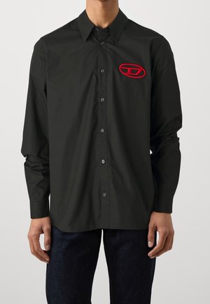 Uomo che indossa una camicia nera con bottoni e maniche lunghe, con un piccolo logo ovale rosso sul petto sinistro e pantaloni scuri.