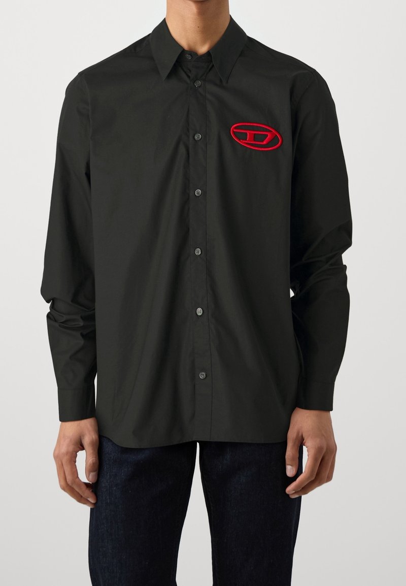 Homme portant une chemise noire à manches longues avec boutons, avec un petit logo ovale rouge sur la poitrine gauche et un pantalon foncé.