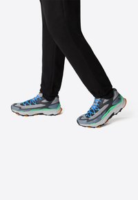 Gray athletic shoes with blue laces, green accents, and textured mesh uppers; thick soles with orange detailing and rugged tread.