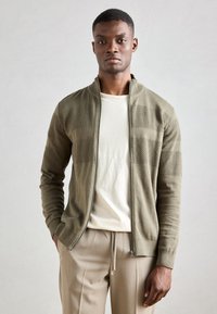 Cardigan verde oliva con zip, caratterizzato da strisce orizzontali testurizzate, tessuto a maglia, polsini e orlo a costine, indossato sopra una t-shirt color beige chiaro.