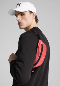 Mochila roja con un zipper y correa negra, que presenta un logo. El modelo lleva un suéter negro y una gorra blanca, con un fondo neutral.