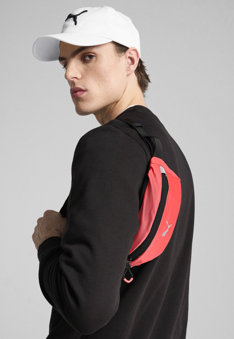Mochila roja con un zipper y correa negra, que presenta un logo. El modelo lleva un suéter negro y una gorra blanca, con un fondo neutral.