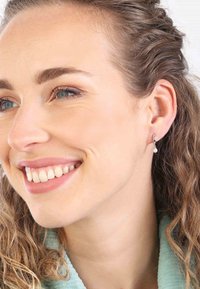 Boucles d'oreilles en argent en forme de hoop avec une petite perle ronde suspendue à chacune. Le modèle a des cheveux ondulés châtain clair et affiche une expression souriante.