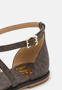 Sandale à motif marron avec une bride à la cheville, munie d'une boucle dorée et d'une semelle intérieure lisse couleur tan embossée du logo "MK". Design classique à bout ouvert.