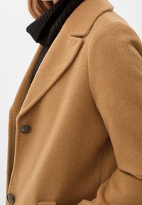 Donna che indossa un cappotto di lana color cammello sopra un maglione a collo alto nero a costine, tagliato all'altezza delle spalle e del petto.