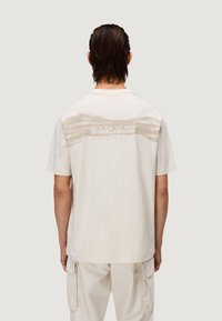T-shirt in cotone crema con una vestibilità rilassata, caratterizzata da una grafica testurizzata sulla parte posteriore in marrone chiaro, che mette in evidenza il marchio "Napapijri" e un design di onde.