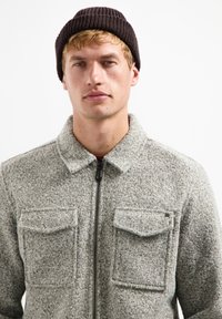 Grijze, gestructureerde zip-up jas met twee borstzakken, een opgerold kraag en een gladde rits. Draagt een donkerbruine gebreide beanie.