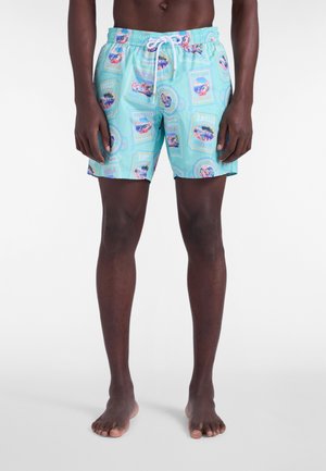 Uomo che indossa pantaloncini da bagno azzurri chiari con stampe di cartoline di viaggio e coulisse bianca, in piedi a piedi nudi su uno sfondo semplice.