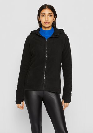 Femme portant un sweat à capuche noir zippé avec poches latérales, un pull à col roulé bleu et des leggings noirs en similicuir, debout devant un fond uni.