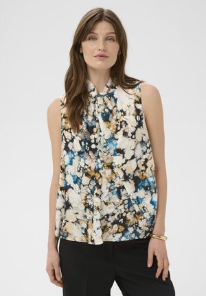 Vrouw draagt een mouwloze blouse met een bloemenpatroon in crème-, blauw- en bruinetinten, gecombineerd met een zwarte broek en een gouden armband.