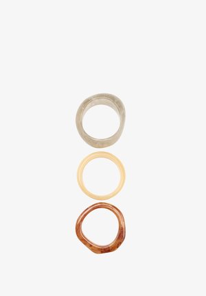 Trzy nałożone na siebie pierścienie: górny w przezroczystym szarym, środkowy w jasnym żółtym, dolny w czerwono-brązowym z teksturowanymi, nieregularnymi krawędziami.