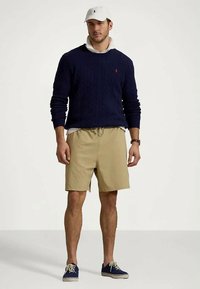 Pulôver azul-marinho em malha de cabos, calções bege com cordão, camiseta branca por baixo e sapatilhas azuis. Um boné com logotipo completa o visual.