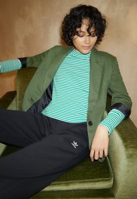 Zaļš šaha raksta žakets virs zaļas un balta svītrainas turtleneck, apvienots ar melniem Adidas sporta biks�ēm, sēžot uz zaļas samta krēsla.