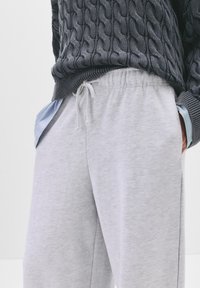 Grå bomulds sweatpants har en elastisk talje med snøre, sidelommer og en afslappet pasform. En blåstribet skjorte kigger frem.