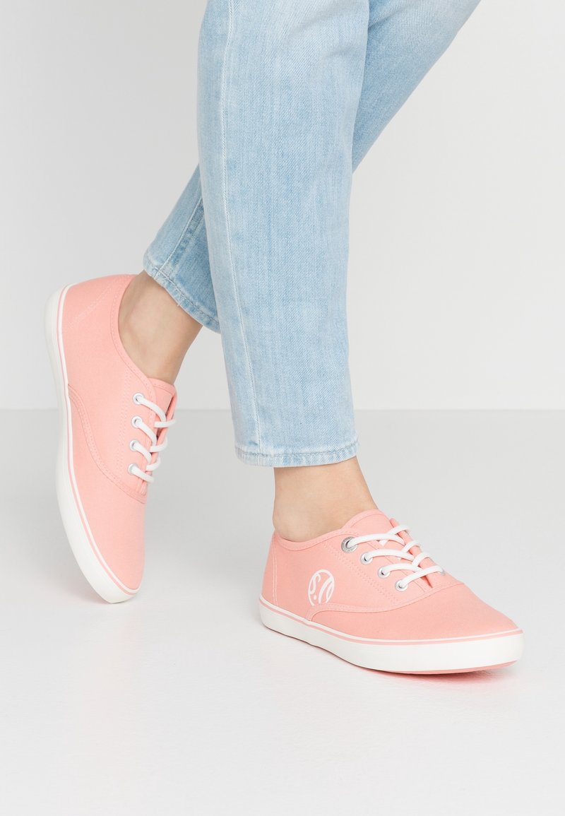 Baskets en toile roses avec lacets blancs et semelles en caoutchouc ; détail du logo sur le côté. Portées avec un jean bleu clair retroussé.