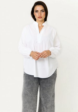 Woman standing against white background wearing a loose white blouse with gathered cuffs and gray acid-wash jeans, hands clasped in front.