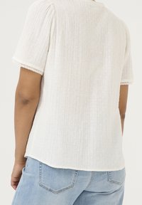 Blusa blanca de manga corta con tela texturizada, con puntos elevados y un borde de encaje en las mangas. Vista trasera muestra un dobladillo redondeado.