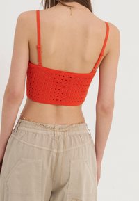 Červený háčkovaný crop top s nastavitelnými ramínky, se strukturovaným designem a otevřeným pleteným vzorem. V kombinaci s béžovými cargo kalhotami a řetízkovým opaskem.