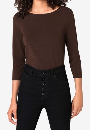Woman wearing a brown three-quarter sleeve top tucked into high-waisted black jeans with five visible front buttons.